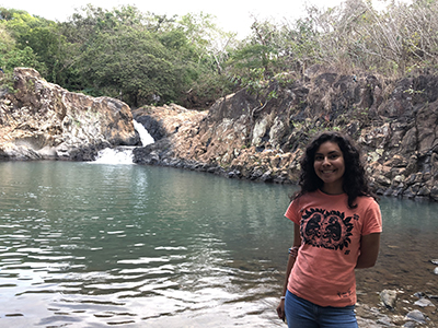 a smiling young college student with long dark hair stands by a rippling pond with a rocky waterfall in the background