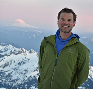 A young bearded man stands on a wintery mountain with Mt Raineer in the far background