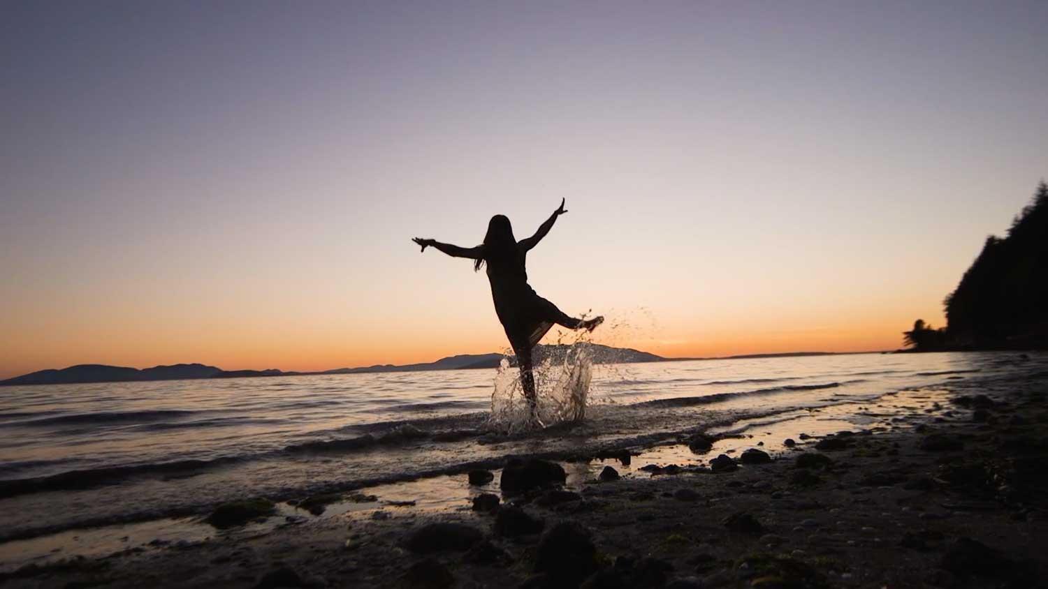 A woman stands on a beach, arms outstretched, playing in the surf during sunset