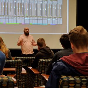 a bearded man in the fornt of class as they lecture a class using a digital audio mixing board on the shown on the movie screen